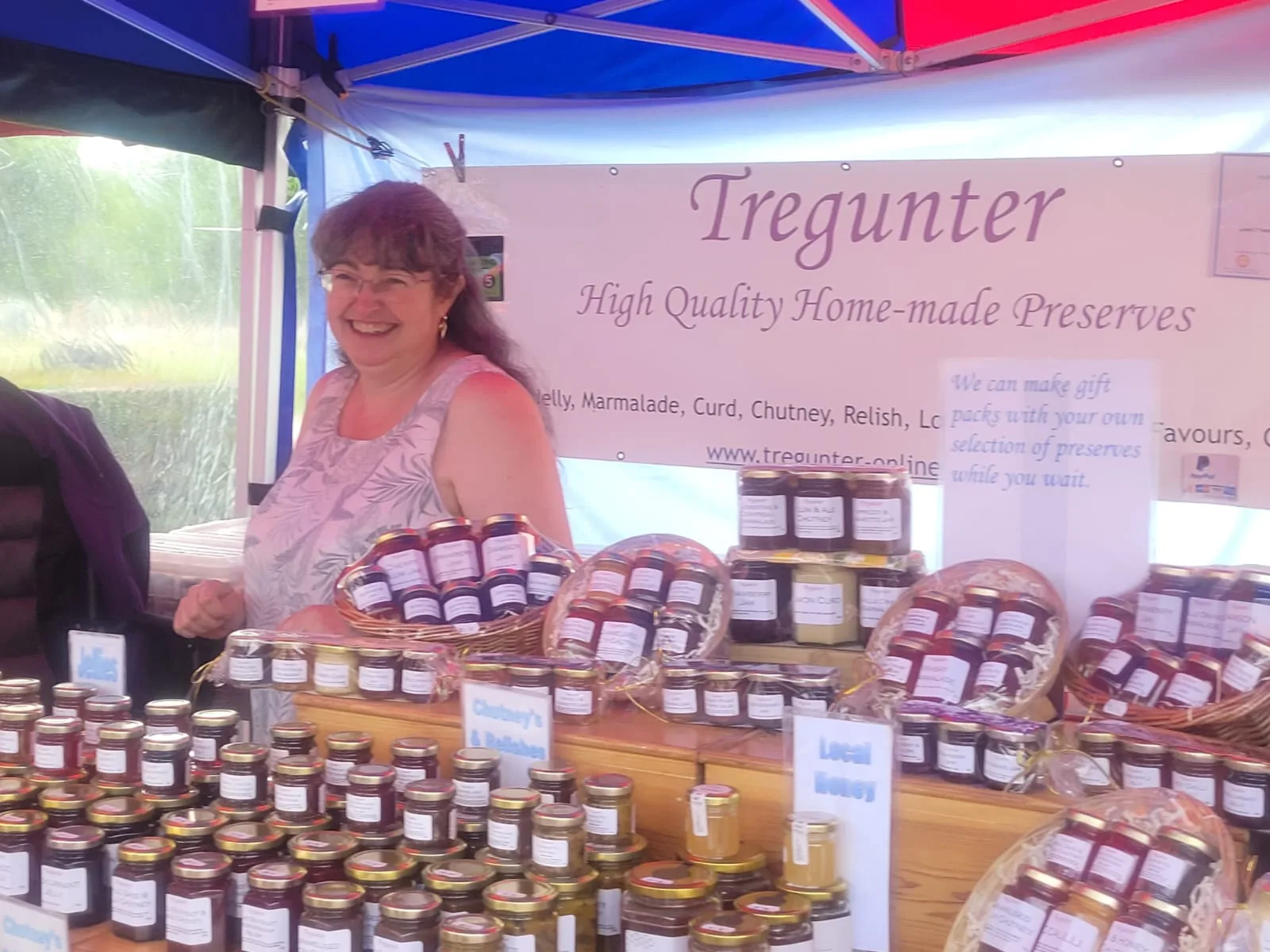 Sarah at her Tregunter market stall — the "High Quality Home-made Preserves" banner overhead, rows of jam jars and gift baskets along the front.